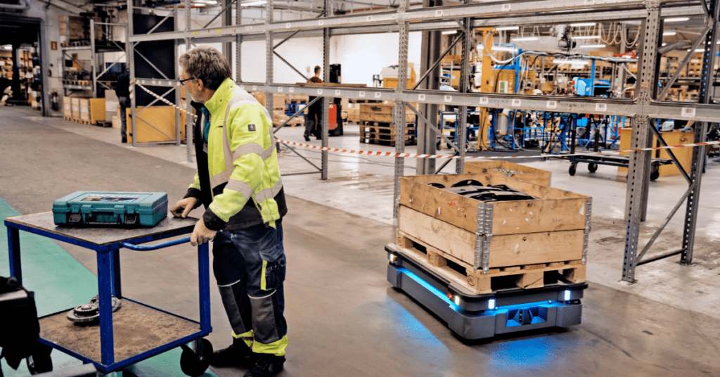 Un operario observa un robot móvil autónomo (AMR) transportando una caja de materiales en una fábrica.