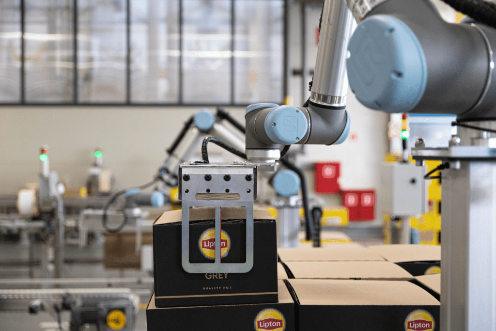 Un entorno industrial con un brazo robótico en primer plano, que parece estar realizando una tarea de empaquetado de cajas de té Lipton.
